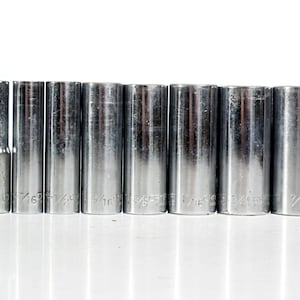 May include: A set of eight cylindrical chrome-plated steel socket wrench attachments arranged in a row. Each socket is engraved with its specific imperial size, ranging from 1/4 inch to 7/8 inch, featuring a polished metallic finish.