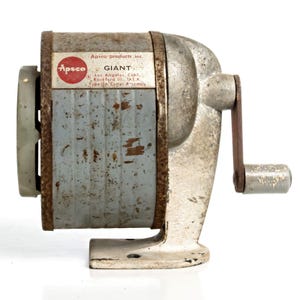 May include: A vintage Apsco Giant pencil sharpener with a weathered grey cylindrical body, a metal crank handle, and a white base. The label reads Apsco Products Inc., Giant, Los Angeles, Calif., Rockford Ill., U.S.A., and Type 3A Cutter Assembly.