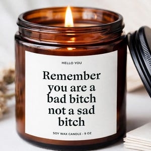 May include: A lit soy wax candle in a brown glass jar with a black lid. The candle has a white label with the text "HELLO YOU Remember you are a bad bitch not a sad bitch." The candle is 9 oz.