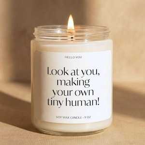 May include: A clear glass jar soy wax candle with a lit wick. The white label features the text HELLO YOU, Look at you, making your own tiny human!, and SOY WAX CANDLE 9 OZ. The candle wax is off-white and the flame is bright yellow and orange.