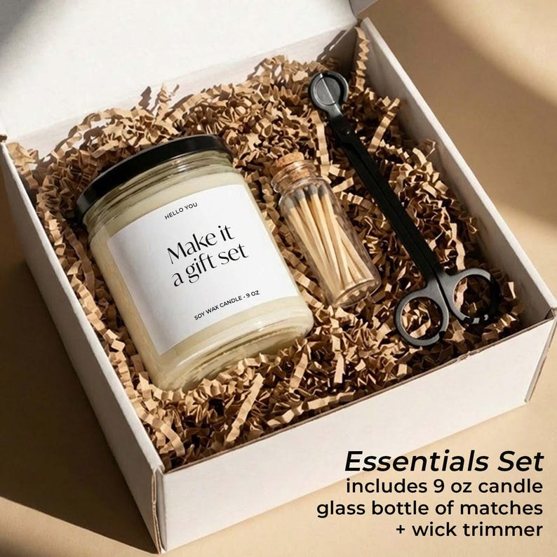 A white box containing a soy wax candle, glass bottle of matches, and a wick trimmer, all wrapped in brown shredded paper.