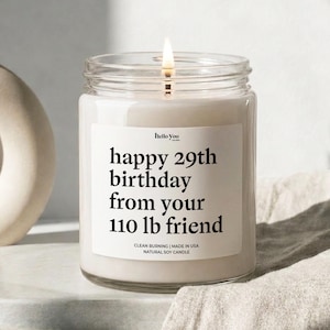 A white candle with a lit wick, placed on a white surface. The candle has a label that reads "happy 29th birthday from your 110 lb friend".