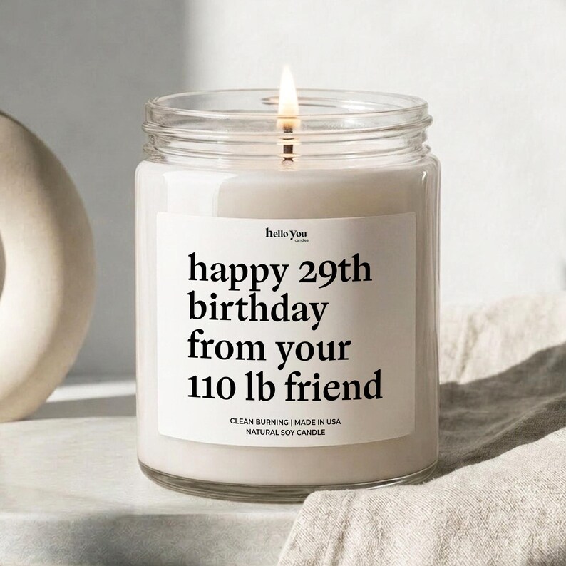 A white candle with a lit wick, placed on a white surface. The candle has a label that reads "happy 29th birthday from your 110 lb friend".