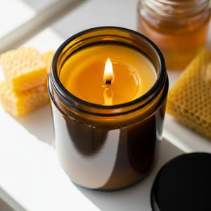 May include: A lit candle in a brown glass jar with a black lid. The candle has a yellow wax and a lit wick. Honeycomb and a jar of honey are in the background. The candle is on a white surface.
