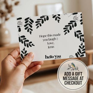 A hand holding a card with a floral design and text, along with a circular sign that reads "ADD A GIFT MESSAGE AT CHECKOUT".