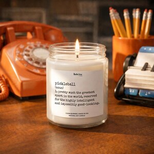 A white candle with a lit flame sits on a wooden desk, surrounded by office supplies such as pencils and a phone.