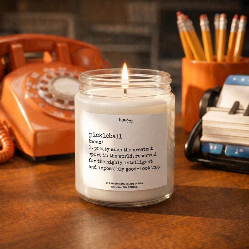 A white candle with a lit flame sits on a wooden desk, surrounded by office supplies such as pencils and a phone.