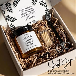 A gift set containing a candle and a pair of scissors, presented in a white box with brown paper bedding. The candle has a label that reads "In loving memory of your forties."