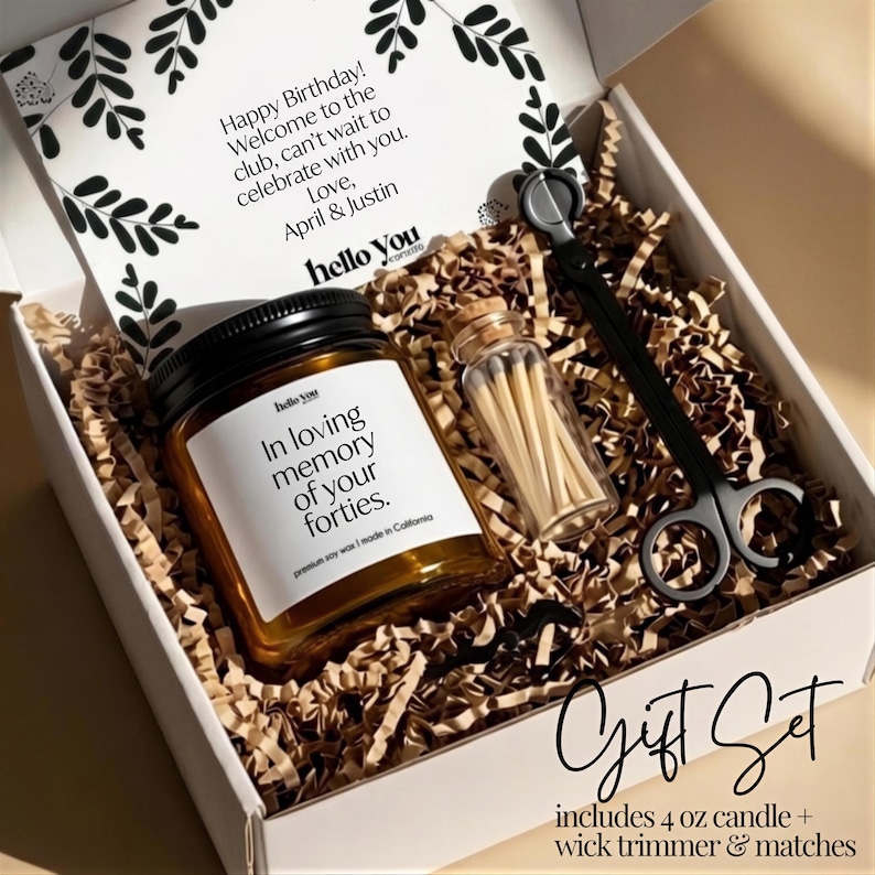 A gift set containing a candle and a pair of scissors, presented in a white box with brown paper bedding. The candle has a label that reads "In loving memory of your forties."