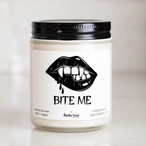 May include: A clear glass candle jar with a black lid containing white wax. The label features a black graphic of lips with fangs and the text BITE ME, along with the text premium soy wax, clean + vegan, and hello you candles, hand-poured in laguna beach, cal.
