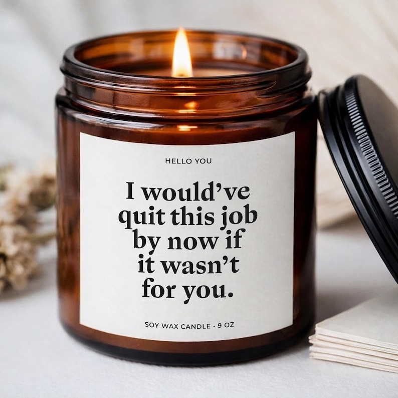 May include: A lit soy wax candle in a brown glass jar with a black lid. The candle has a white label with the text "HELLO YOU" and "I would've quit this job by now if it wasn't for you." The candle is 9 oz.