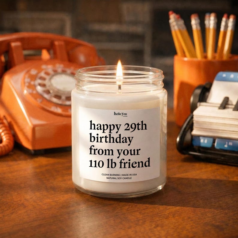 A white candle with a message on it, placed on a wooden desk next to a telephone and a cup of pencils.