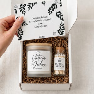 A hand is holding a white box containing a candle and a matchbox, with a card inside the box.