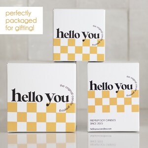 May include: Three white boxes with a black and yellow checkered pattern and the text "hello you" in black. The boxes are stacked on top of each other. The top box has the text "the original candle greeting" in black. The bottom box has the text "PREMIUM SOY CANDLES SINCE 2015 helloyoucandles.com" in black.