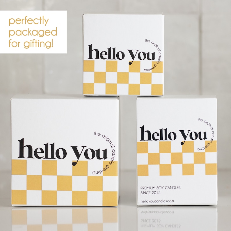 May include: Three white boxes with a black and yellow checkered pattern and the text "hello you" in black. The boxes are stacked on top of each other. The top box has the text "the original candle greeting" in black. The bottom box has the text "PREMIUM SOY CANDLES SINCE 2015 helloyoucandles.com" in black.