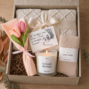 Puede incluir: Una caja de regalo que contiene una manta texturizada de color crema atada con una cinta, un ramo de tres tulipanes de ganchillo, una vela con la etiqueta No Words Just Hugs y un par de calcetines beige con la etiqueta cozy socks y una tarjeta de Winnie.