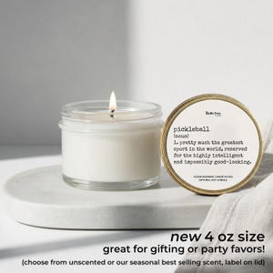 A white candle in a glass jar with a label next to it, and a white cloth or towel on a white surface.