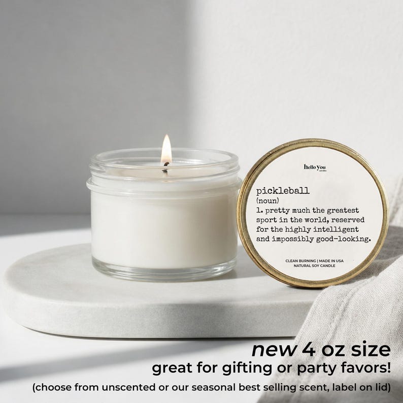 A white candle in a glass jar with a label next to it, and a white cloth or towel on a white surface.