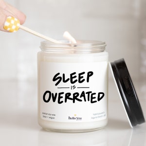 May include: A white candle with a black label that reads "SLEEP IS OVERRATED". The candle is made with premium soy wax and is clean and vegan. The candle is hand-poured in Laguna Beach, California.