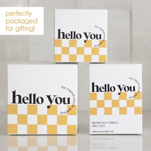 Puede incluir: Tres cajas blancas con un patr&oacute;n de cuadros negros y amarillos y el texto "hello you" en negro. Las cajas est&aacute;n apiladas una encima de la otra. La caja superior tiene el texto "the original candle greeting" en negro. La caja inferior tiene el texto "PREMIUM SOY CANDLES SINCE 2015 helloyoucandles.com" en negro.