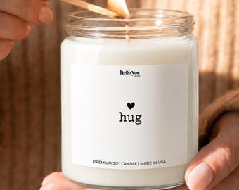May include: A clear glass jar candle filled with white wax, featuring a white label with the text hello you, a small black heart icon, the word hug, and the text PREMIUM SOY CANDLE | MADE IN USA. A wooden matchstick lights the candle wick.