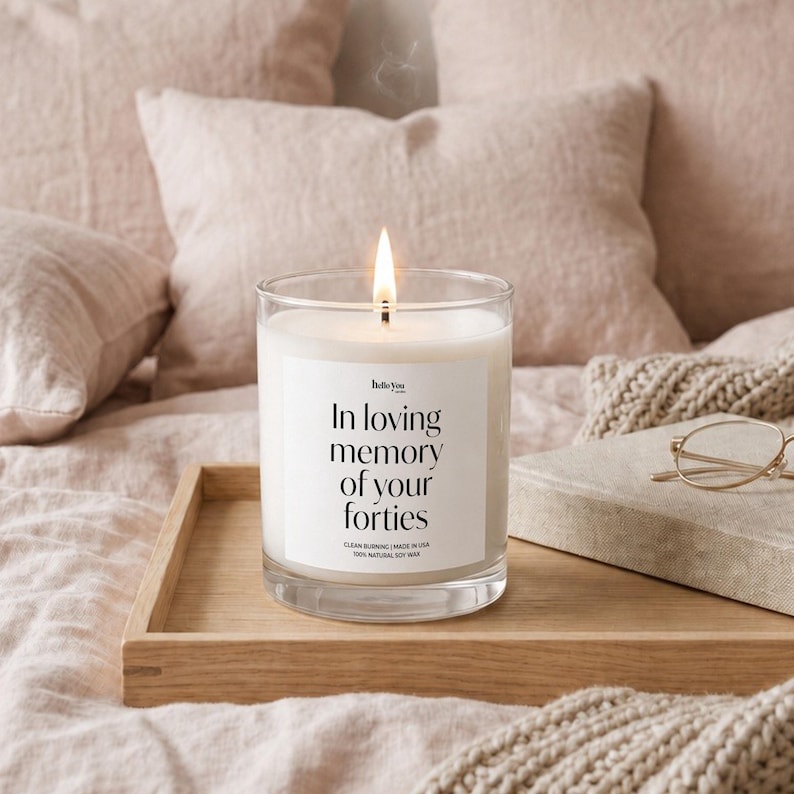 A lit candle on a wooden tray, with a pair of glasses and a book nearby, sits on a bed with a beige blanket.