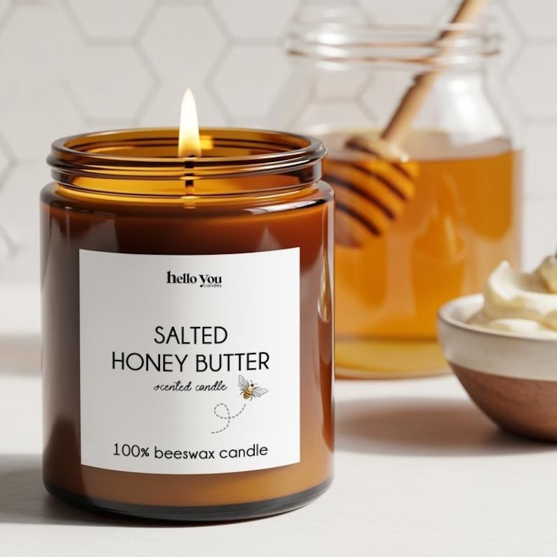 May include: A lit, brown glass jar candle with a white label that reads "Salted Honey Butter" and "100% beeswax candle." A jar of honey and a small bowl of cream are in the background. The candle has a bee graphic.