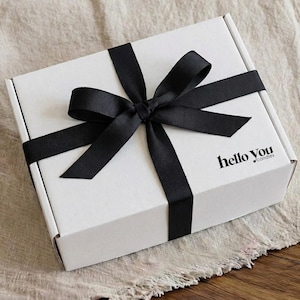 A white gift box with a black ribbon tied around it, sitting on a beige cloth.