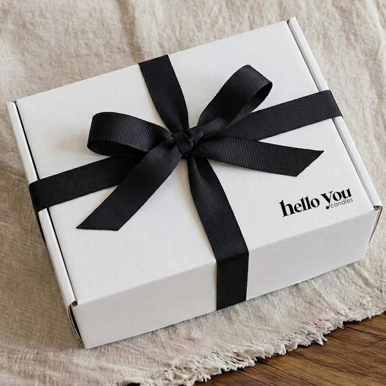 A white gift box with a black ribbon tied around it, sitting on a beige cloth.