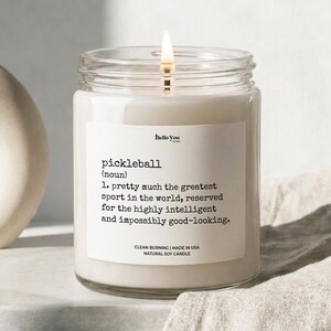 A white candle with a lit wick, placed on a surface with a beige cloth. The candle has a label that provides a definition of pickleball, which is a sport that is considered to be pretty much the greatest sport in the world, reserved for the highly intelligent and impossible to look at.