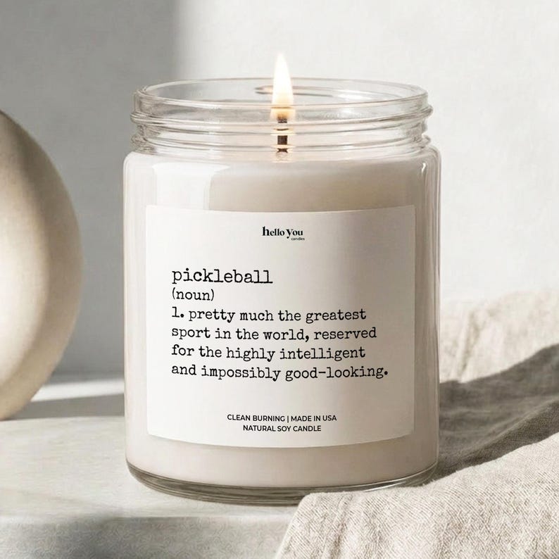 A white candle with a lit wick, placed on a surface with a beige cloth. The candle has a label that provides a definition of pickleball, which is a sport that is considered to be pretty much the greatest sport in the world, reserved for the highly intelligent and impossible to look at.