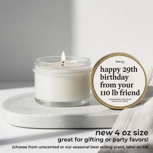 A white candle in a glass jar with a label next to it, and a white cloth or napkin on a white surface.