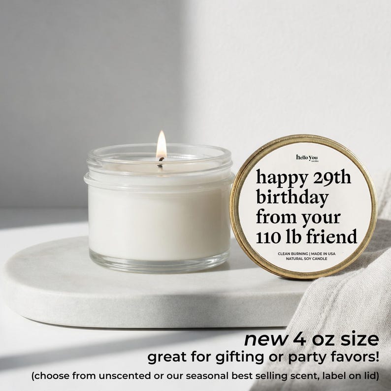A white candle in a glass jar with a label next to it, and a white cloth or napkin on a white surface.