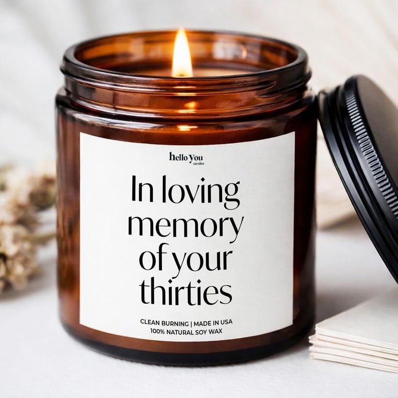 Może przedstawiać: Zapalona świeca w bursztynowym szklanym słoiku z czarną metalową pokrywką. Biała etykieta zawiera tekst "hello you candles", "In loving memory of your thirties", "CLEAN BURNING | MADE IN USA" oraz "100% NATURAL SOY WAX" czcionką szeryfową.
