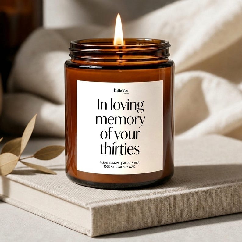 Może przedstawiać: Zapalona świeca w brązowym szklanym słoiku z białą etykietą zawierającą tekst "hello you", "In loving memory of your thirties", "CLEAN BURNING | MADE IN USA" oraz "100% NATURAL SOY WAX" wydrukowany czarną czcionką szeryfową i bezszeryfową.