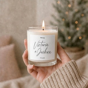 A hand holding a lit candle with a label that says "Hello You" and "Victoria + Joshua" on it, with a blurred background of a Christmas tree and a couch.