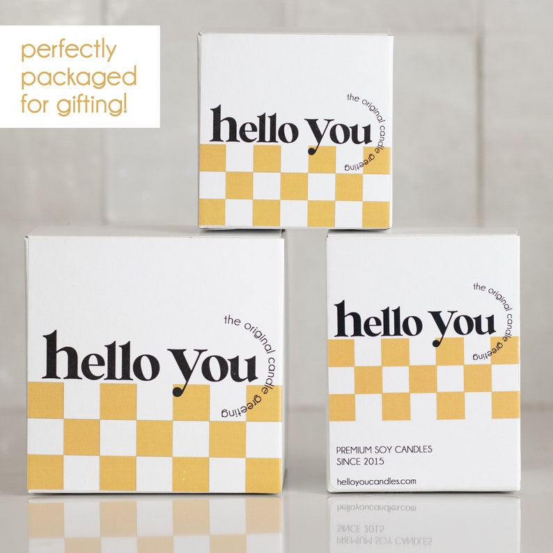 May include: Three white boxes with a black and yellow checkered pattern and the text "hello you" in black. The boxes are stacked on top of each other. The top box has the text "the original candle greeting" in black. The bottom box has the text "PREMIUM SOY CANDLES SINCE 2015 helloyoucandles.com" in black.