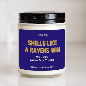 a white candle with a black lid, placed on a white surface. The candle has a label that reads "Hello You Smells Like a Ravens Win My Lucky Game Day Candle kick ass candles since 2015".