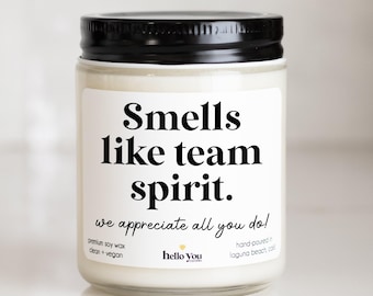 May include: A clear glass candle jar with a black lid containing white wax. The label features the black text Smells like team spirit. we appreciate all you do! premium soy wax clean + vegan hello you candles hand-poured in laguna beach, cal.