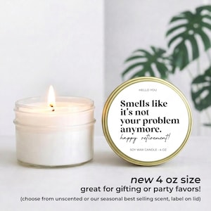 May include: A lit white soy wax candle in a clear glass jar next to a gold-rimmed tin with the text "Smells like it's not your problem anymore. Happy retirement!" The tin is labeled "SOY WAX CANDLE - 4 OZ".