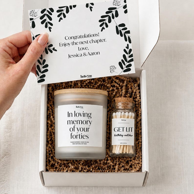 A hand is holding a white box containing a candle and a jar of matches, with a card inside the box.