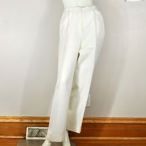 May include: White, high-waisted pants with a straight leg, displayed on a mannequin. The pants have a classic design with a button and zipper closure. The mannequin is positioned against a white wall with framed artwork and a thermostat.