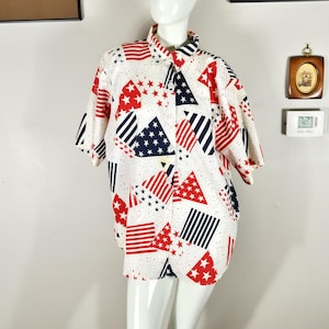 Puede incluir: Camisa blanca con botones con un patrón patriótico de triángulos, estrellas y rayas rojas, blancas y azules. La camisa de manga corta se exhibe en un maniquí. La camisa es de estilo casual.