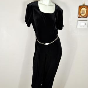 Könnte beinhalten: Schwarzer Samt-Jumpsuit mit rundem Ausschnitt und kurzen Ärmeln. Ein silberner Ketten-Gürtel mit ovalem Mittelstück betont die Taille. Der Jumpsuit wird auf einer Schaufensterpuppe präsentiert.