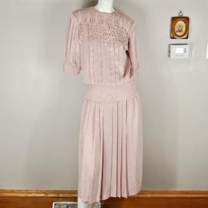 Puede incluir: Vestido rosa pálido vintage sobre un maniquí. El vestido presenta un canesú de encaje detallado, mangas cortas y una falda plisada. La tela tiene un sutil estampado floral. El vestido se exhibe en una habitación bien iluminada.