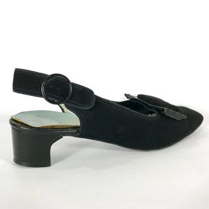 Puede incluir: Zapato negro con correa trasera y tacón pequeño. El zapato presenta un cierre de hebilla en la correa del tobillo y un lazo decorativo en la puntera. El zapato tiene una puntera redondeada y un tacón de bloque pequeño.