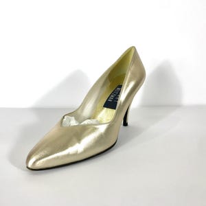 May include: A gold-colored high heel pump with a pointed toe. The shoe has a stiletto heel and a low-cut vamp. The brand name "Stuart Weitzman" is visible on the insole. The shoe is set against a white background.