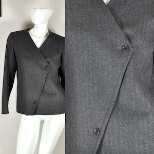 May include: A gray blazer with a V-neck and long sleeves. The blazer features a wrap-style front with two buttons for closure. The fabric appears to be a medium-weight material, suitable for cooler weather. The blazer is displayed on a mannequin.