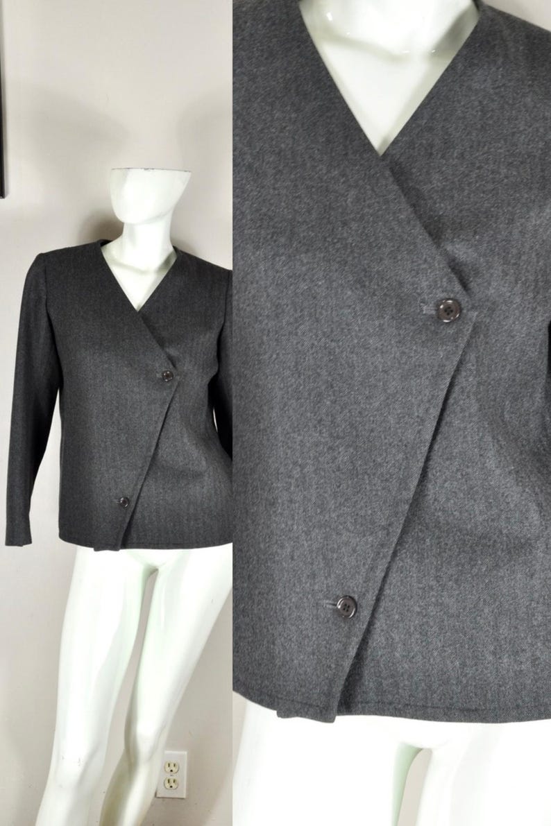 May include: A gray blazer with a V-neck and long sleeves. The blazer features a wrap-style front with two buttons for closure. The fabric appears to be a medium-weight material, suitable for cooler weather. The blazer is displayed on a mannequin.