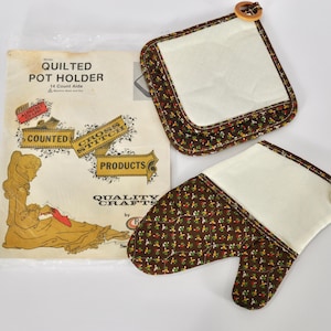 Op de afbeelding: Een set gewatteerde pannenlappen en een ovenwant met een bloemenpatroon in bruin, rood en groen op een crèmekleurige achtergrond. De pannenlap en want hebben houten ringaccenten. De verpakking leest "Quilted Pot Holder".